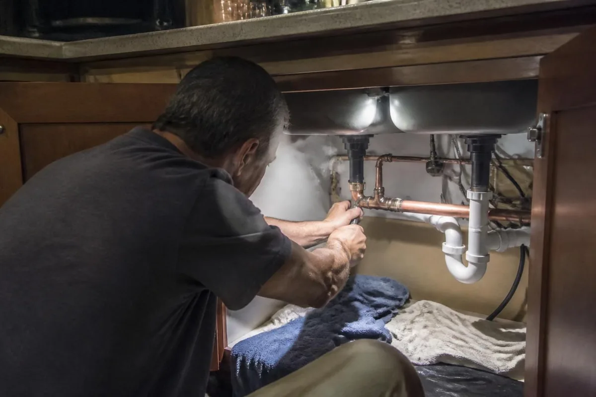 Plumber repairing a split copper pipe under a kitchen sink at night, towels on the floor, emergency plumber in Raleigh, NC